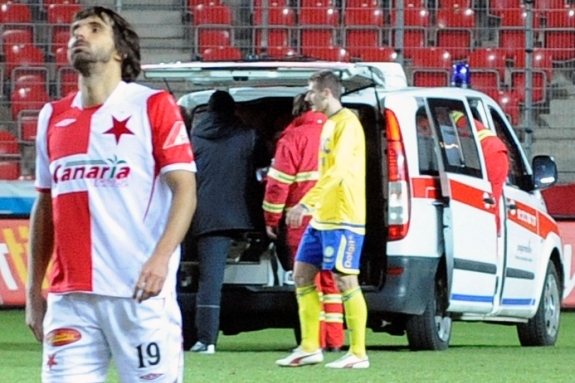 Fotbalista Slavie Matej Krajčík, za ním odváží sanitka Rista Naumova.