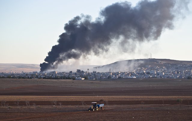 Traktor jede po poli, v pozadí hoří část města Kobani po útoku amerického letectva na pozice Islámského státu. 
