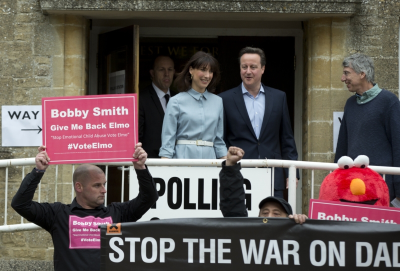 David Cameron s manželkou vycházejí z volební místnosti, která je obložená protestujícími.