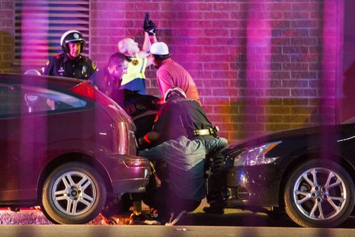 Policista kryje své spolupracovníky, kteří odnášejí zraněného kolegu z dosahu střelby neznámých útočníků.