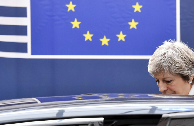 Theresa Mayová nastupuje do auta. V pozadí vlajka EU. 