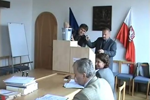 Notářka (sedící u stolu) se směje nestandardnímu průběhu losování.